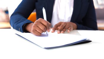 Businesswoman signing contract. African American business woman sitting at table in office, holding pen and writing in document. Legal expertise concept