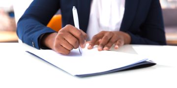 Businesswoman signing contract. African American business woman sitting at table in office, holding pen and writing in document. Legal expertise concept