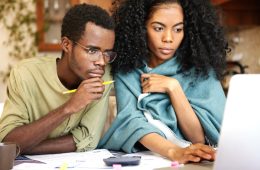 Young African wife and husband with many debts doing paperwork together, analyzing expenses, planning family budget and calculating bills, sitting at kitchen table with laptop, calculator and papers
